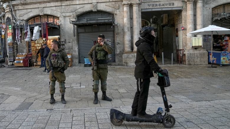 Two Israeli soldiers wearing uniforms stand guard in Jerusalem's Old City last month as a  person wearing a black helmet rides by on a scooter.