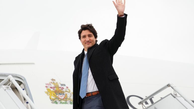 Justin Trudeau, wearing a jacket and tie, waves in front of an airplane with Canada's coat of arms on it.