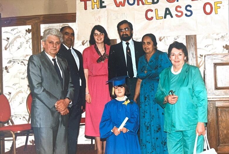 A smiling child in graduation robes is surrounded by six adults. 