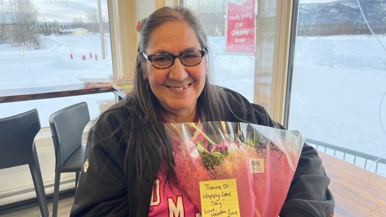 A smiling woman sits in a room holding a wrapped bouquet of flowers.