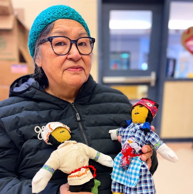 An Elder sits on a chair holding two dolls. One has a plaid dress and embroidered red hat, the other has a white hat and cloth jacket. 