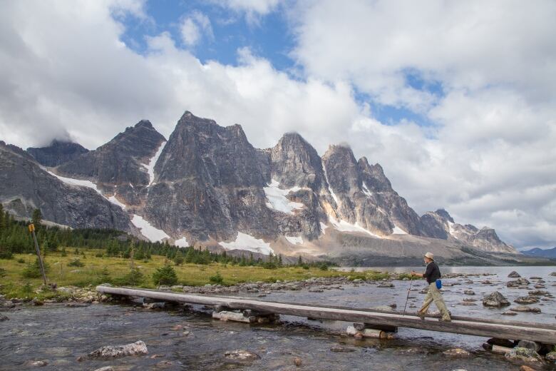 A man crosses a wooden foot bridge that spans a creek in the foreground, with a series of mountain peaks called The Ramparts in the background.