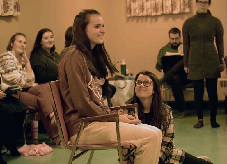 Young woman with long brown hair sits on chair, another young woman with long brown hair and glasses sits at her feet. Other young women sit on chairs in background.