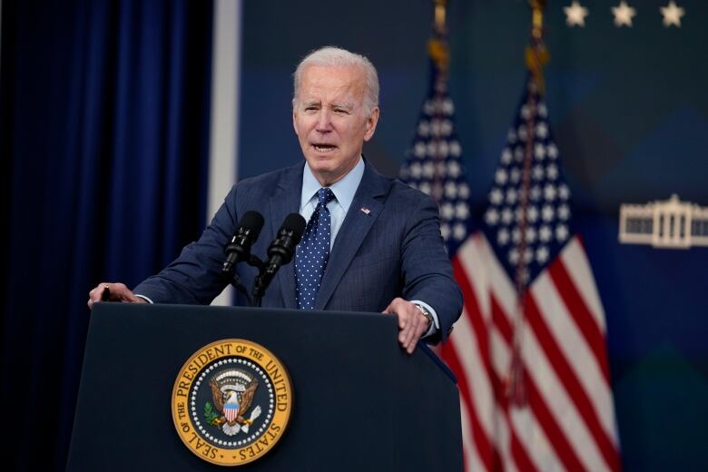 An older man in a suit stands at a podium that says Seal of the President of the United States. There are U.S. flags behind him.