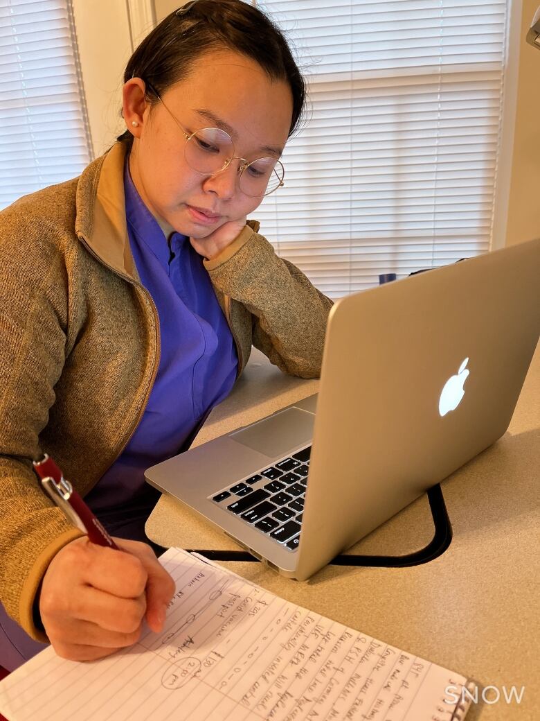 A person sitting in front of a laptop computer writes in a notebook.