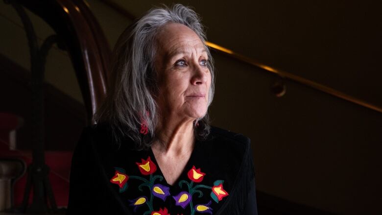 A grey-haired woman in a black top with embroidered flowers looks to her left.