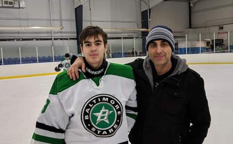 An older man in a black coat and toque stands at a hockey rink with his arm around a younger man. They're on the ice, and the younger man (his son) is in a Baltimore Stars jersey.