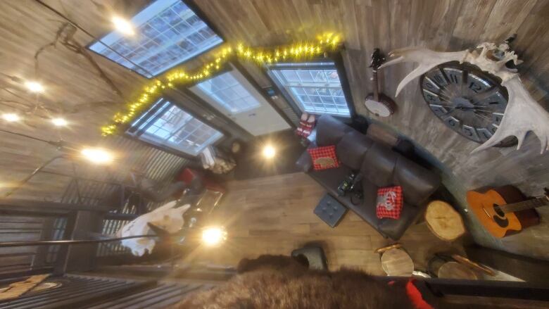 A view looking down from a loft railing showing a couch, fireplace and stools below. A bear skin run, antlers, an acoustic guitar and banjo hang on the wall. The area is lit with a variety of hanging lights.