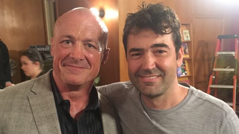 A man with a bald head, grey suit jacket and black shirt poses with a man with brown hair and a grey T-shirt on a TV filming set.