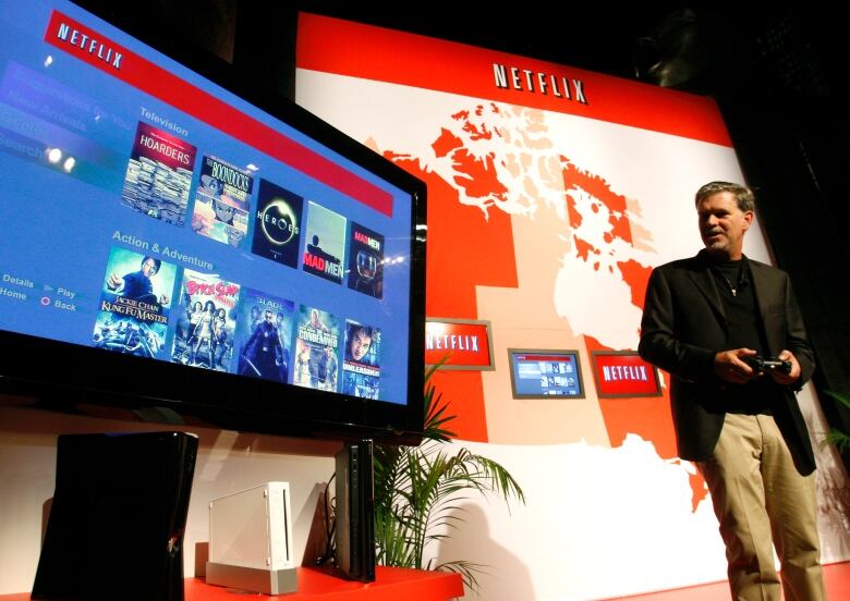 Man with brown hair, wearing a black suit and khaki pants, stands on a conference stage, looking at a large TV screen showing Netflix shows.