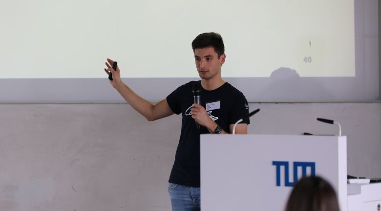 A man gestures to a projected image while addressing a classroom.