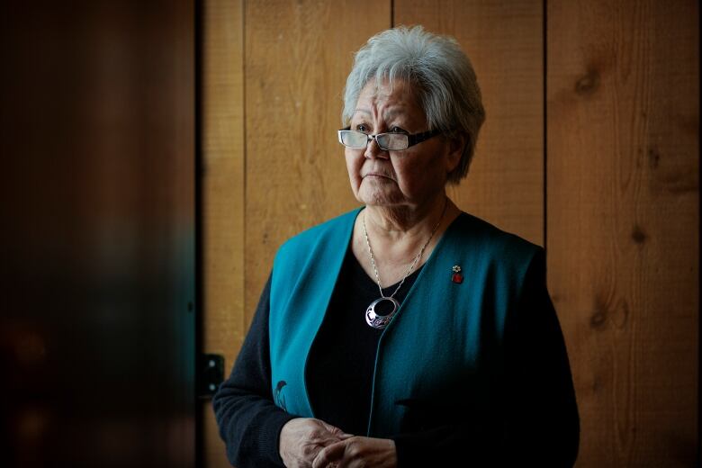 An elderly woman stands for a portrait in front of a wood wall.