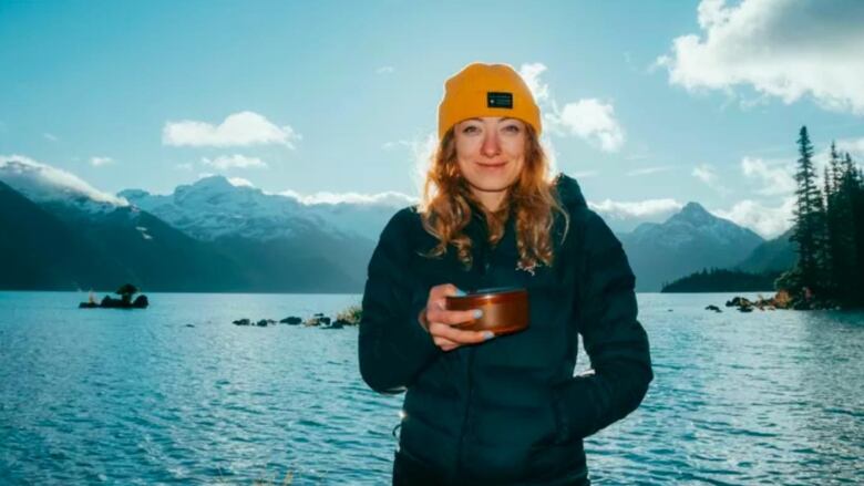 A woman with red hair and a yellow toque smiles for a photo on a sunny day. A lake and mountains are in the background.