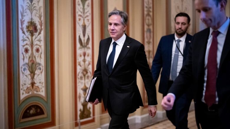 A politician holding a binder walks along a hallway with two other men.