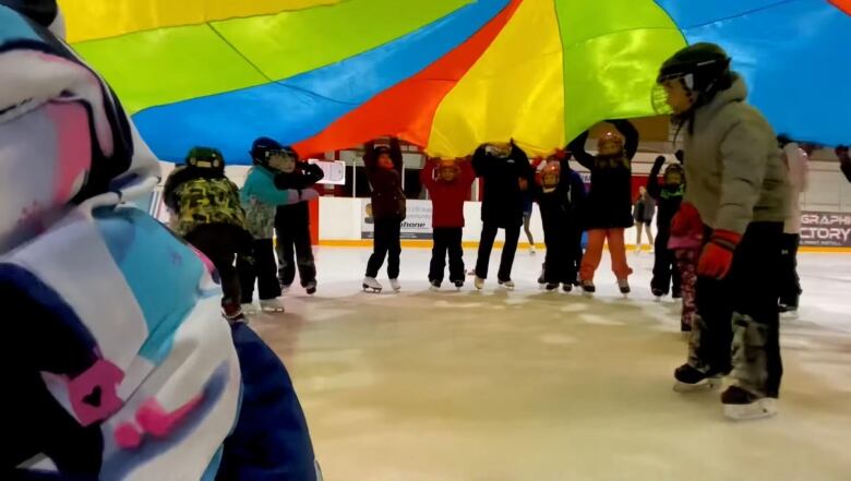 Kids are seen on skates, standing in a circle, and holding a colourful fabric above their heads as others skate through.