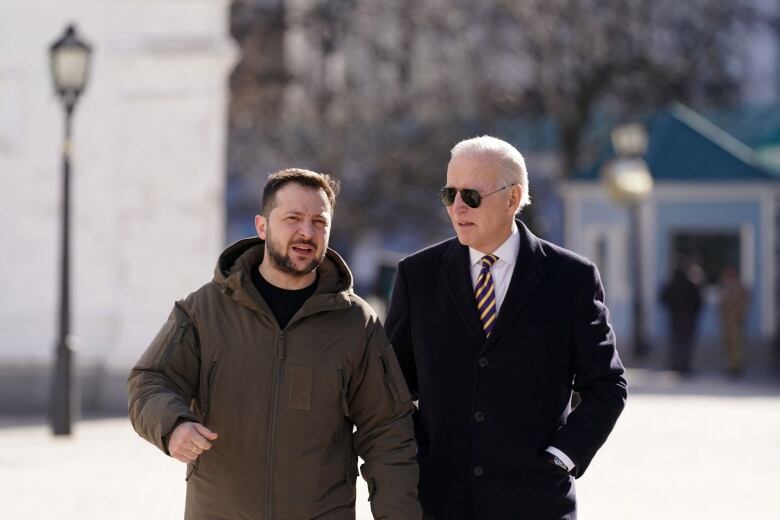 U.S. President Joe Biden wearing suit and tie walks next to Ukrainian President Volodymyr Zelenskyy wbearing brown jacket on sunny street