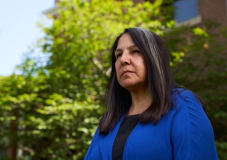 A woman with long black hair and grey streaks wearing a blue jacket looks away from the camera with a trees in the background.