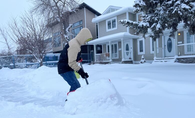 A man in a winter coat and hood shovels snow from a sidewalk.