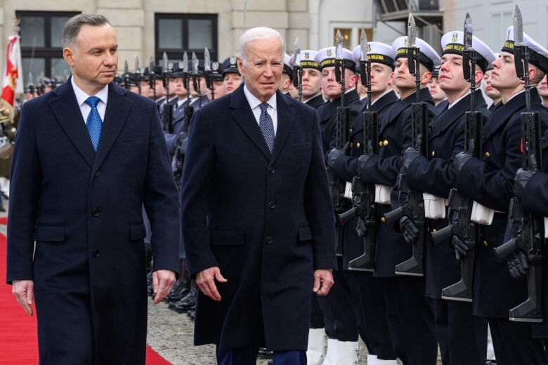 Two men in coats are shown walking along a row of troops.