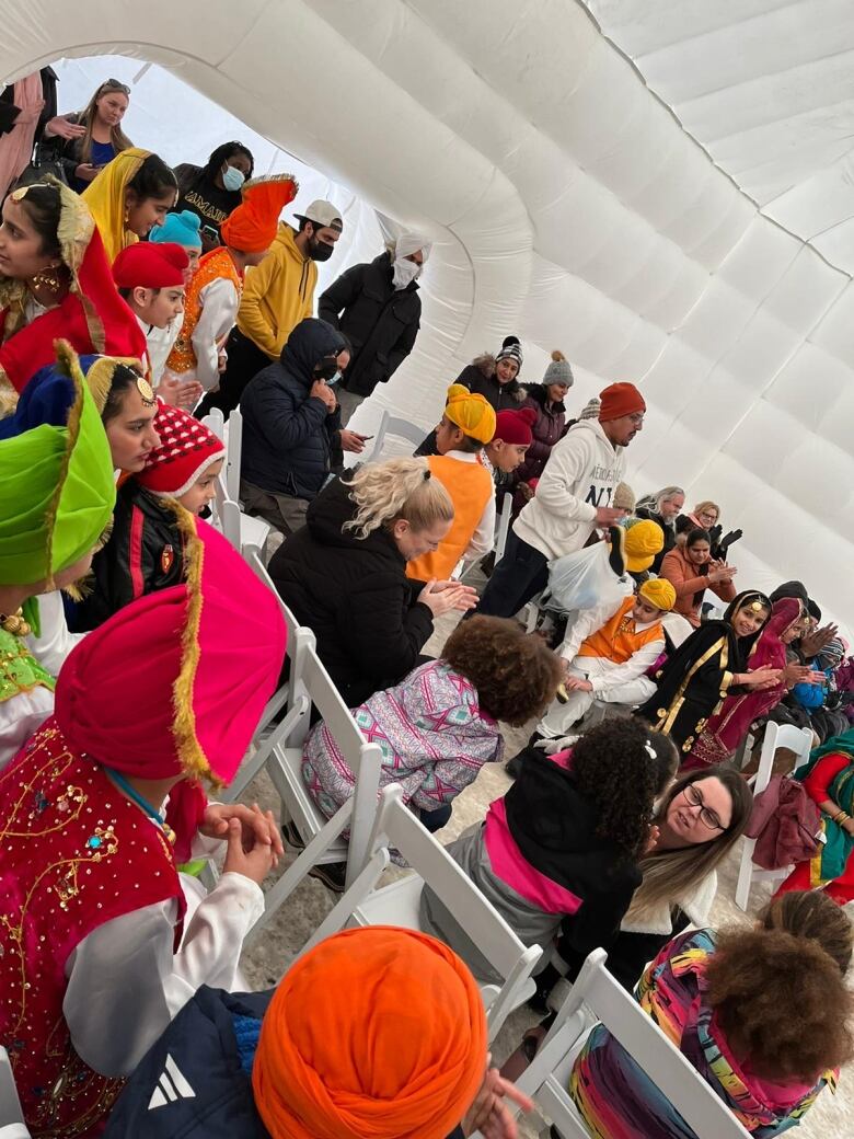 A diverse crowd sits on white chairs under a white tent, listening to performances at the IRAL Winter Festival. 