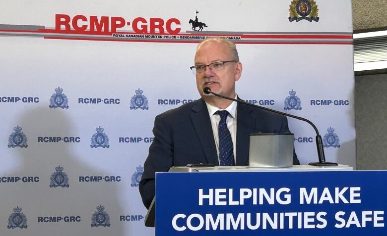 A balding man with glasses looks out from behind a podium, which has a poster that says 