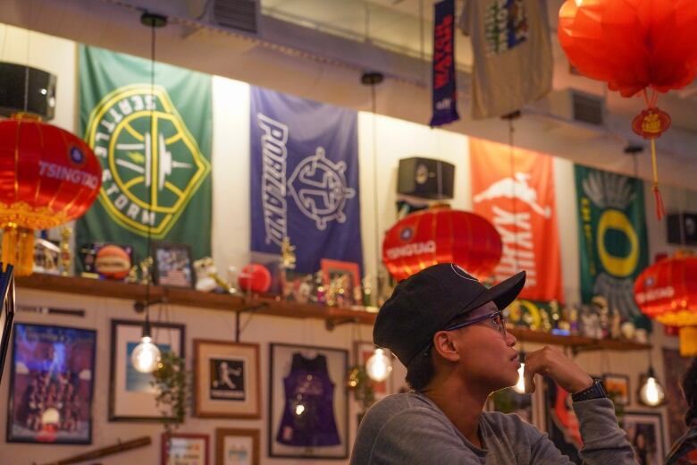 A woman with short hair and glasses wears a baseball hat while watching a television in a bar decorated with memorabilia from women's sports teams. 