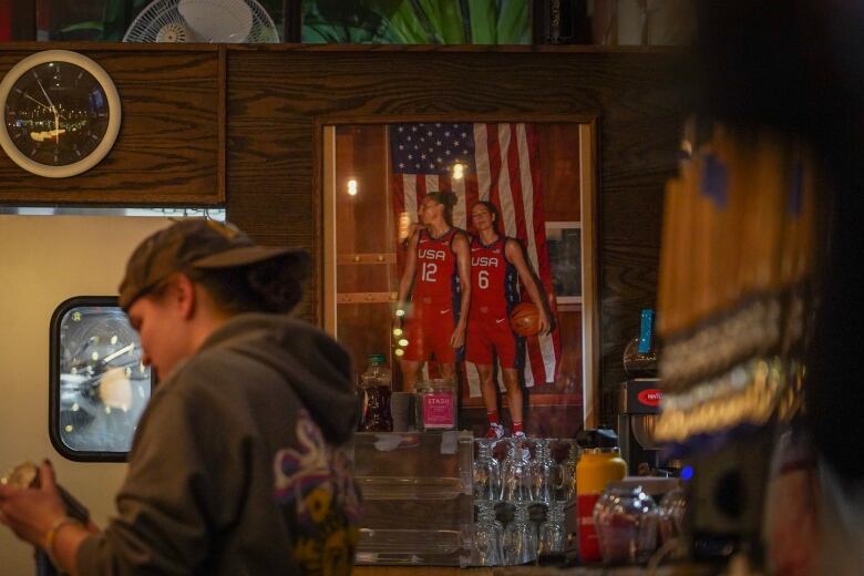 A staff member wearing a hoodie and backwards baseball hat works behind a bar decorated with women's sports memorabilia, including a framed photo of Team U.S.A. women's basketball players Diana Taurasi and Sue Bird.