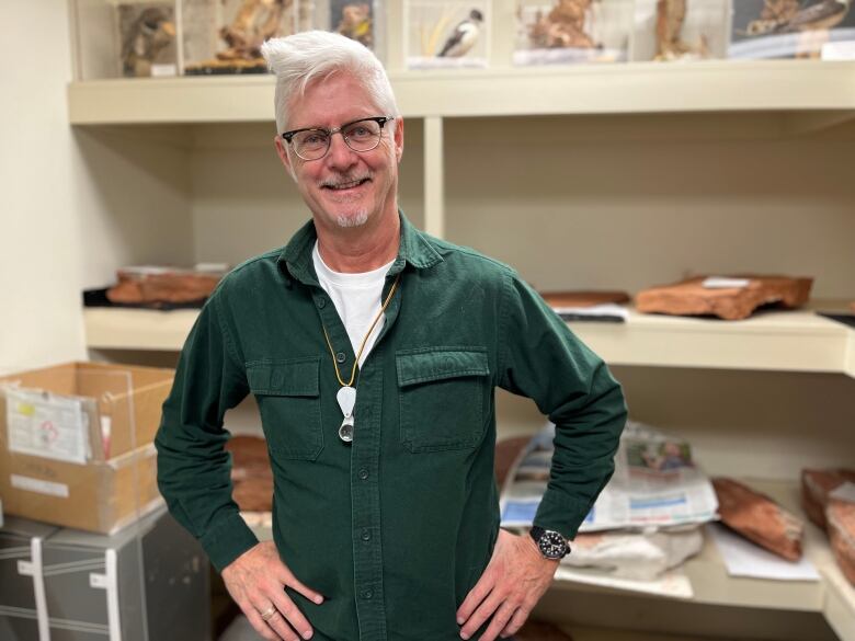 Man with grey hair and glasses wearing dark green shirt over white T-shirt stands, hands on hips, smiling at camera.