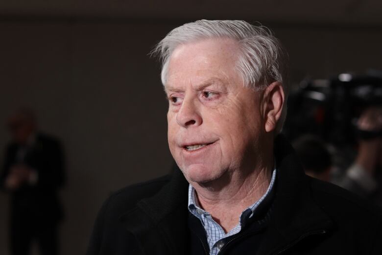 A man with white hair wearing a black sweater over a button up dress shirt showing looking to his right. 