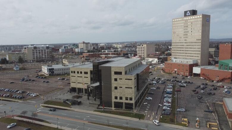 A five storey building clad in stone and glass shown surrounding by parking lots with other downtown Moncton buildings in the background.