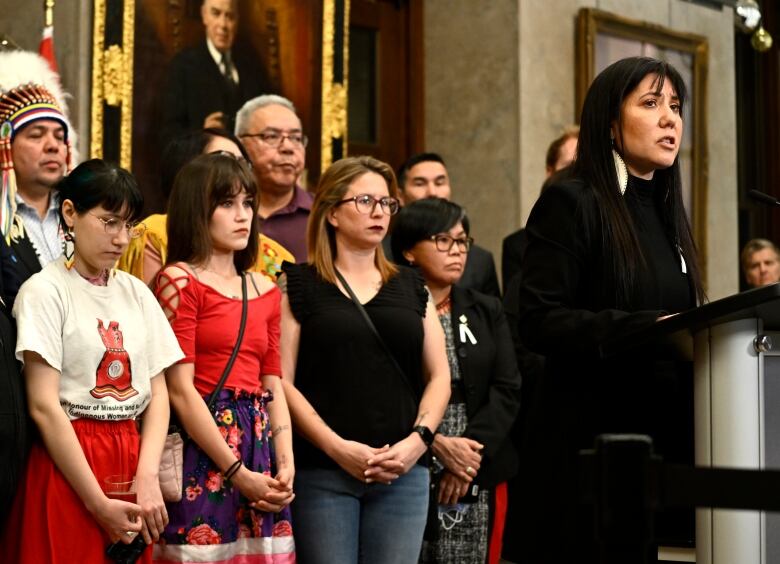 A woman with long dark hair stands at a podium in front of a group of other people who look solemn, some with their heads bowed.