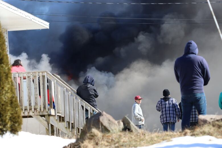 People stand on steps to a house and on sidewalk and watch as dark smoke billows from a nearby house fire.