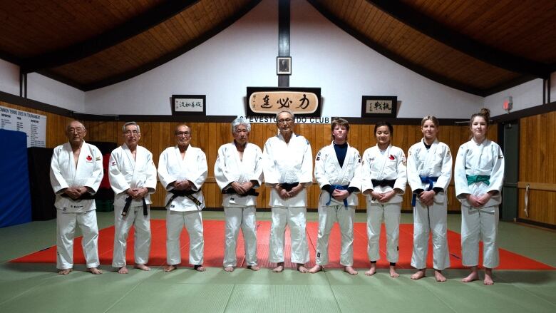 NIne people in white judo attire pose for the camera in a gym with a pagoda-style roof and a Japanese sign in the back.