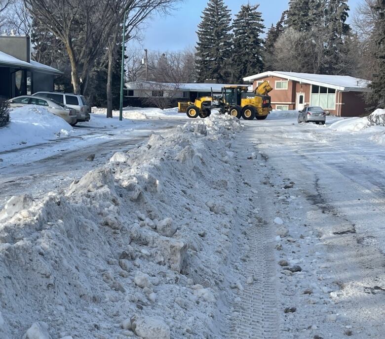 A snow plow in the background is shown working to clear a large piled row of snow, along a neighbourhood street, in the foreground. 