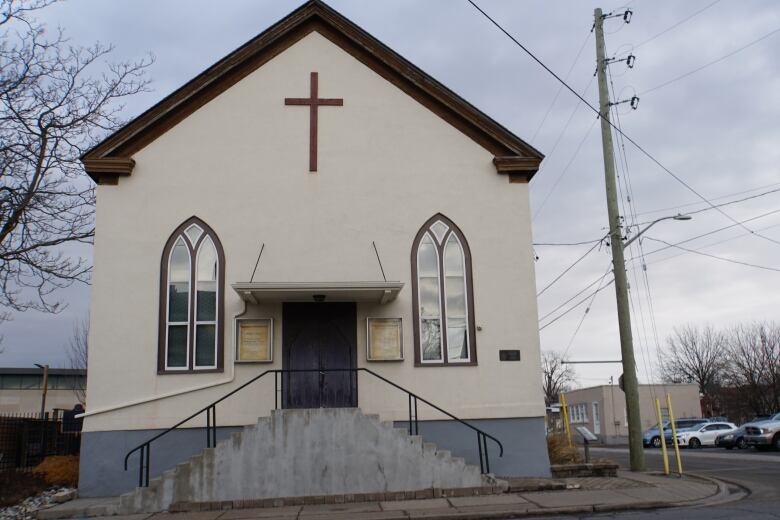 A small church with stairs out front and a big brown cross on the front of it can be seen from the road. 