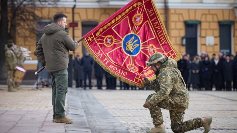 A leader holds a large flag while a soldier kneeling in front of him holds the other end of it.