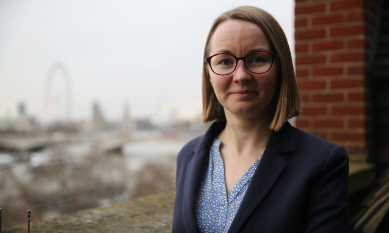 A woman with blond hair who's wearing glasses and a navy blazer stands on a balcony.