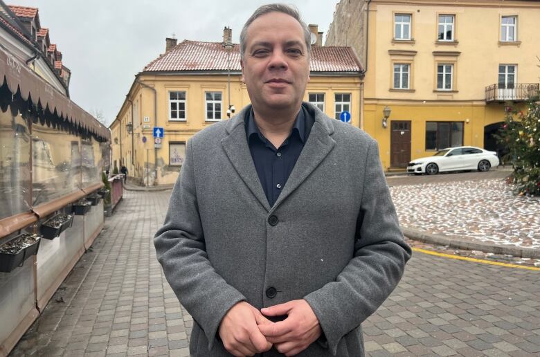 A man wearing a grey overcoat stands on a cobblestoned street.