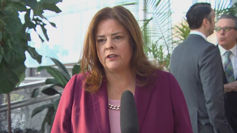 A woman with long brown hair wears a burgundy-coloured jacket and stands behind a microphone.