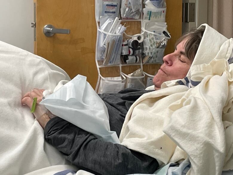 Heather Smith lies in a hospital bed with her eyes closed. She is covered with blankets, with one wrapped around her head. She has an IV in her hand.