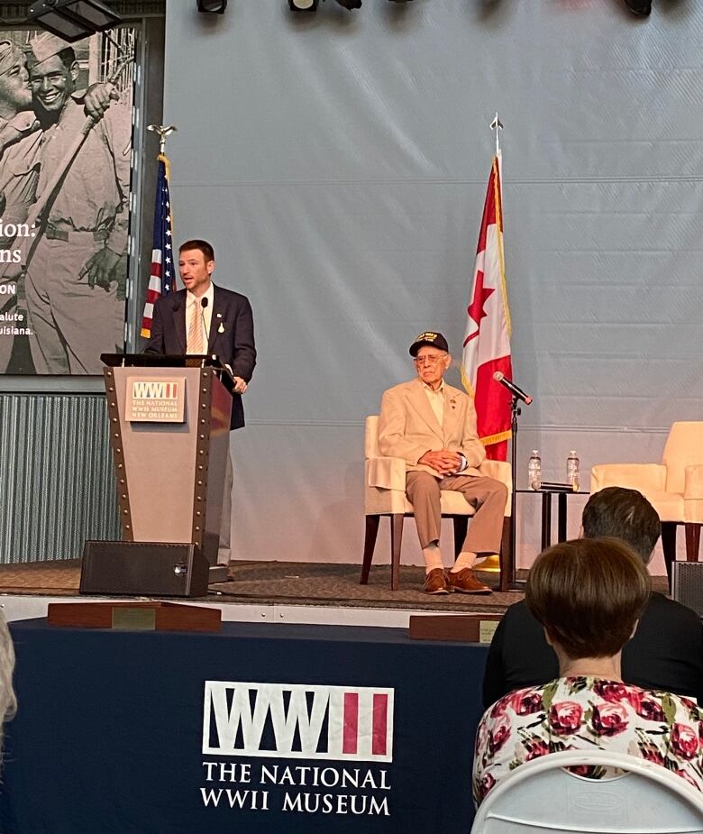 A man speaks to a crowd from behind a podium on a stage. Behind him is seated an elderly veteran.