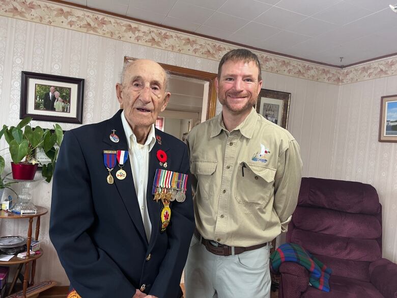 An elderly veteran, with several military medals on his jacket is pictured with a second younger man standing inside the living room of a small home.