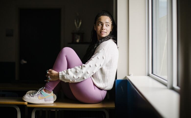 A teacher in pink leggings sits on a desk by a window.