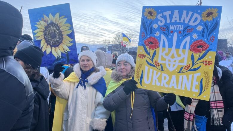 Two women in parkas hold signs in a crowd. One woman has a sign with a picture of a sunflower, while the other is a blue and yellow sign that says 