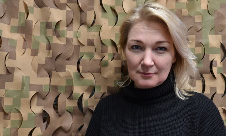 A woman with blond hair poses in front of a camouflage-coloured background. 