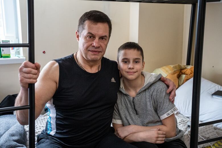 A father poses for a picture with his arm around his son, both seated on a bed. 