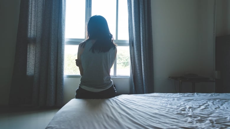 A woman sits on the edge of a bed with one arm folded. She's looking out an open window as light casts a shadow over her and creates a silhouette.