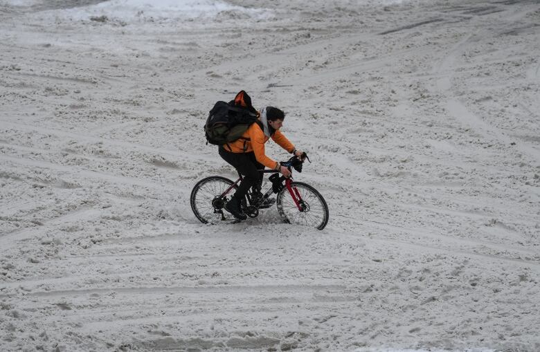 A cyclist in an orange coat navigates through the snow on an unplowed street.