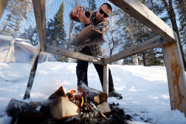 A man seasons a fish on a grill over a wood fire.