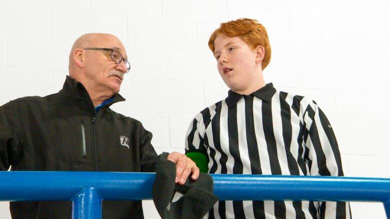 A middle-aged man wearing a black jacket talks with a teenage boy wearing a black and white striped referee's shirt.